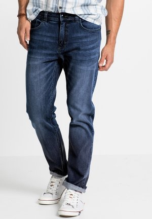 Jeans in denim blu scuro con vestibilità dritta, dotati di tasche frontali, leggere sfumature e orli risvoltati, abbinati a sneaker alte bianche.