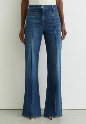 Jean en denim bleu taille haute avec couture sur le devant, jambes droites larges et fermeture par boutons, porté avec des chaussures pointues à motifs.