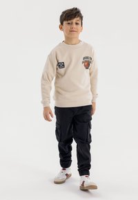 Felpa beige con grafica "Brooklyn Basketball" e toppe, abbinata a pantaloni cargo neri e sneakers bianche con dettagli marroni.