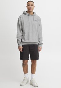 Grauer Kapuzenpullover mit vorderer Tasche, kombiniert mit dunklen Shorts und weißen Sneakers. Das Modell hat eine kurze Frisur und sichtbare Tattoos.