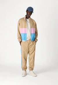 Bež brezrokavnik z vzorcem in rožnatimi ter modrimi paneli, kombiniran z usklajenimi hlačami. Oblečeno s svetlo majico in modro kapo, obuto z belimi športnimi copati.