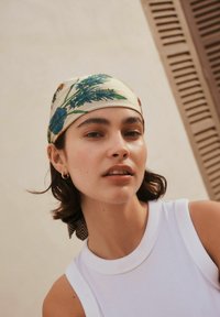 Pañuelo de seda con base beige claro que presenta patrones florales en azul y rojo. Modelo con un top blanco sin mangas, peinado con el cabello suelto.