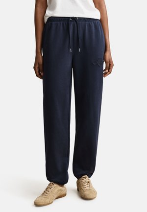 Personne portant un pantalon de sport bleu marine avec taille à cordon et des baskets en daim beige, debout devant un fond blanc uni.