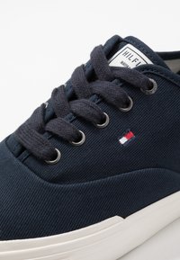 Marineblaue Canvas-Sneaker mit dicken, dunklen Schnürsenkeln, metallenen Ösen, einer weißen Gummisohle und einem kleinen Logo in Rot- und Weißtönen.