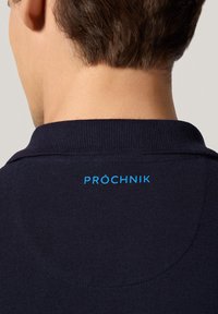 Granatowa koszulka polo o fakturowanym materiale, z niebieskim logo "PRÓCHNIK" na plecach kołnierza oraz klasycznym kroju kołnierza.