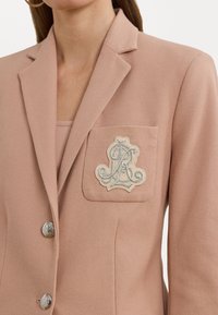 Blazer couleur pêche en tissu texturé, fermeture à un bouton, et détail de poche brodé avec un motif de lettre.