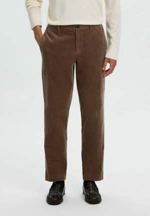 Homme portant un pantalon en velours côtelé marron, des mocassins noirs et un pull crème à manches longues, debout les mains près des poches.