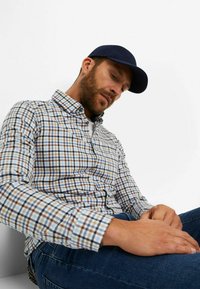 Ein Hemd mit Knopfleiste in einem mehrfarbigen Karomuster mit braunen, blauen und weißen Farbtönen; kombiniert mit dunkelblauen Jeans und einer marineblauen Mütze.