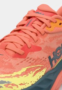 Coral running shoe with textured mesh upper, contrasting yellow and blue accents, and flat laces. Features prominent branding on the side.
