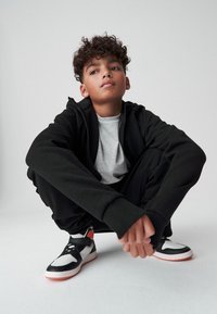 Felpa nera con zip, t-shirt grigia e pantaloni jogger neri. Sneakers bianche e nere con accenti arancioni. Capelli ricci e sfondo neutro.