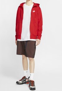 Röd zip-up huvtröja med framfickor, vit logotyp. Buren över en vit t-shirt, ihop med mörka rutiga shorts och svarta/orangea sportskor.