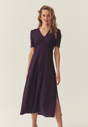 Moteris su bangytais šviesiais plaukais, vilkinti purpurinę midi ilgio suknelę trumpomis rankovėmis ir šoniniu plyšiu, stovinti priešais paprastą šviesią foną.