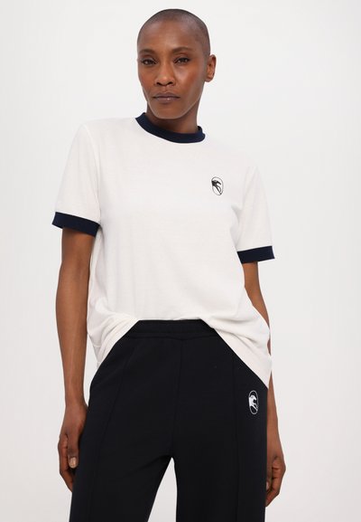 Cream short-sleeve t-shirt with navy ribbed collar and sleeves, featuring a small logo on the chest, paired with dark jogger pants.