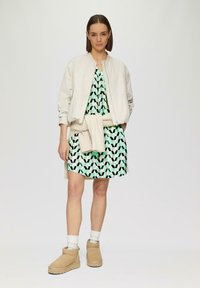 Helle beige Bomberjacke über einem grün gemusterten Kleid mit geometrischem Muster, kombiniert mit hellbraunen Knöchelstiefeln und weißen Socken, Stehend vor einem schlichten Hintergrund.