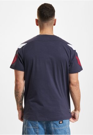 Mann med kort mørkt hår og tatovert arm iført en mørkeblå t-skjorte med røde og hvite skulderdetaljer og blå denimshorts, sett bakfra.