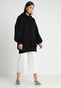 Crni oversized dukserica s prednjim džepom, kapuljačom s vezicom i rebrastim rukavima; u kombinaciji s bijelim prugastim culottes hlačama i crnim kariranim slip-on cipelama.