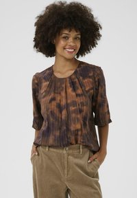 Blouse en tie-dye marron et violet avec des manches trois-quarts, présentant des plis et un col arrondi, associée à un pantalon en velours côtelé beige.