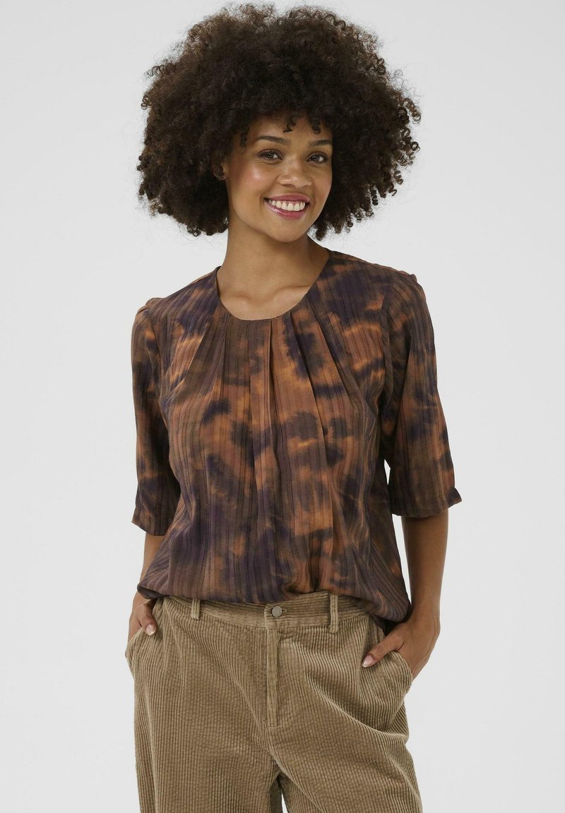 Blouse en tie-dye marron et violet avec des manches trois-quarts, présentant des plis et un col arrondi, associée à un pantalon en velours côtelé beige.