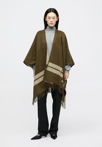 Brown wool poncho with a light gray striped pattern, featuring fringe hems. Worn over a gray turtleneck and black trousers.