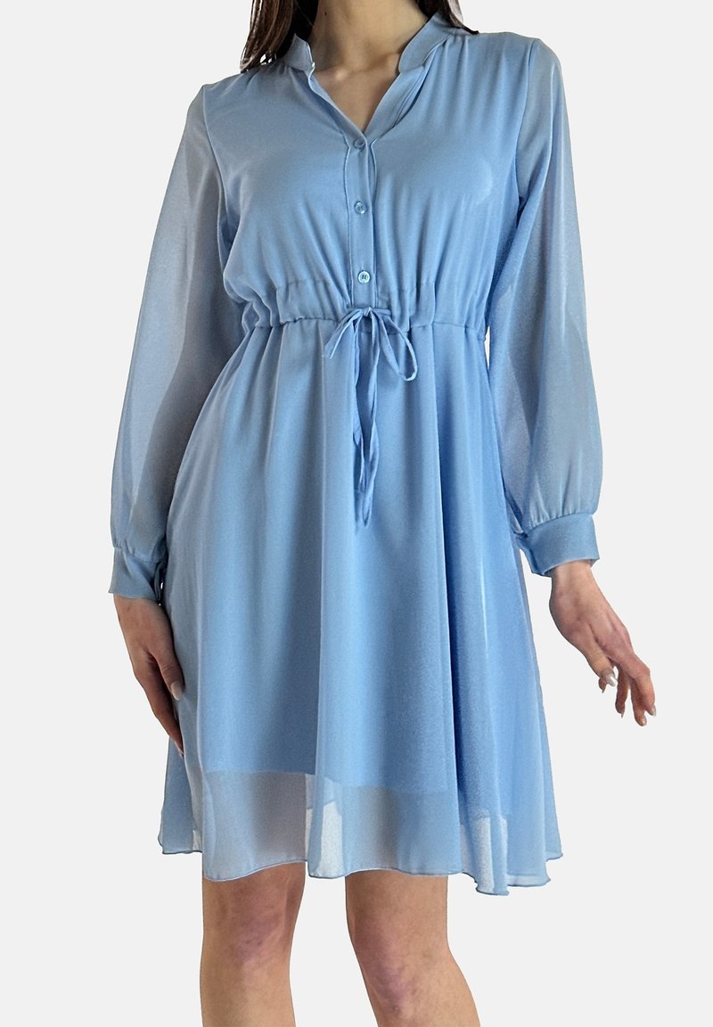 Femme portant une robe bleu clair à manches longues et longueur genou, avec une taille ajustable par cordon et un col à boutons, se tenant devant un fond uni.