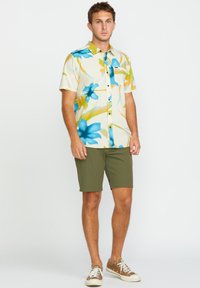 Chemise à manches courtes avec un motif floral bleu et jaune sur un fond crème, associée à un short vert olive et des baskets.