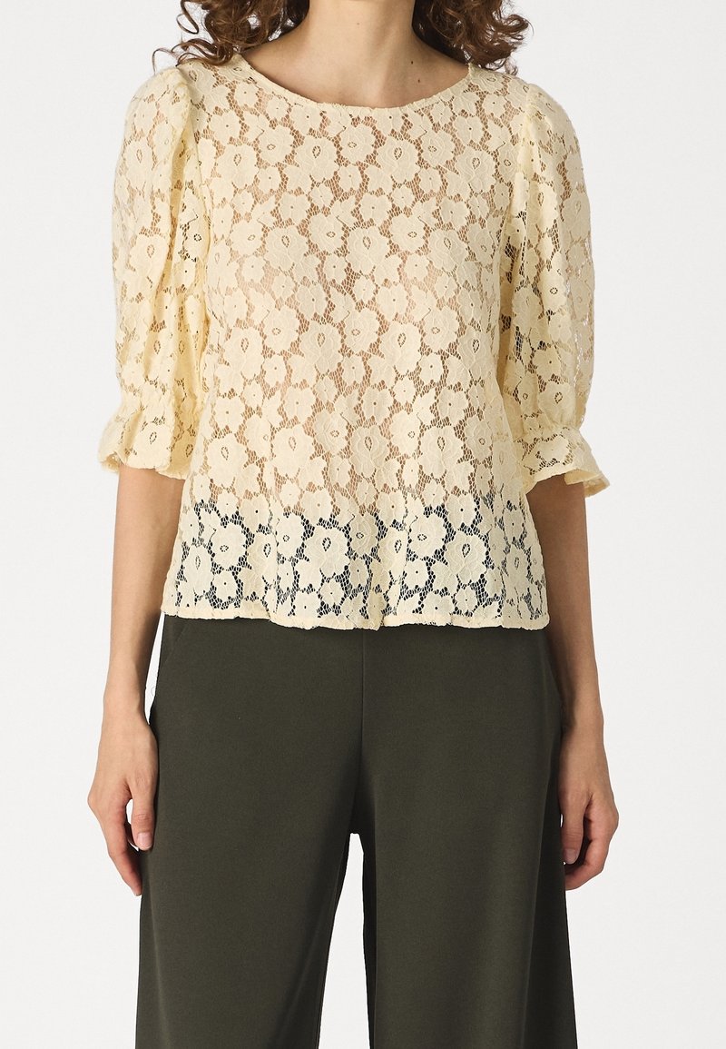 Femme portant un chemisier en dentelle crème à motifs floraux avec des manches bouffantes trois-quarts et un pantalon large kaki foncé.