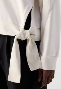 Crème-kleurige sweatshirt met een geknoopte detail aan de zijkant en geribbelde manchetten, aangevuld met een zwarte broek op de achtergrond.
