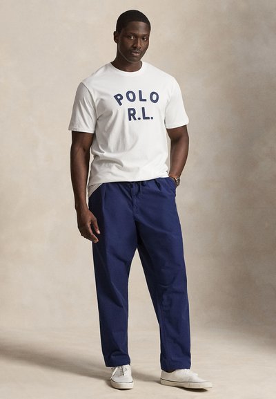 Άνδρας όρθιος φορώντας λευκό μπλουζάκι "POLO R.L.", μπλε παντελόνι, λευκά αθλητικά παπούτσια και ρολόι χειρός σε ουδέτερο μπεζ φόντο.