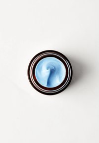 Blå krem i et rundt, mørkebrunt glass med klar lokk. Teksturen ser glatt og kremet ut, med et lite virvelmønster på toppen.