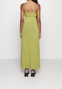 Robe maxi longue couleur vert olive avec bretelles spaghetti ajustables, détails volants, design à étages et tissu doux. Portée avec des sandales noires à semelles épaisses.