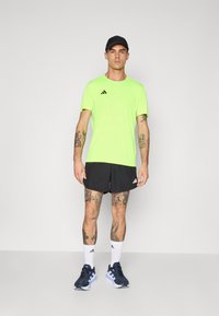 Man in een neon groene Adidas T-shirt, zwarte shorts, witte Adidas sokken, marineblauwe hardloopschoenen en een zwarte pet, staand tegen een effen achtergrond.