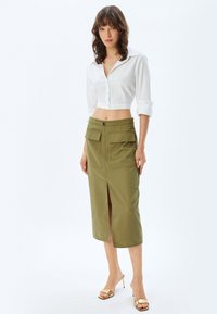 Chemise blanche à manches retroussées, coupée, associée à une jupe haute en olive avec des poches avant et une fente à l'avant.