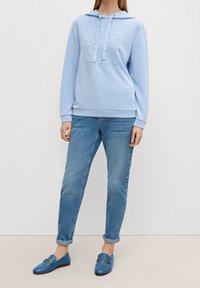 Felpa azzurra con cappuccio e tasca frontale, abbinata a jeans blu arrotolati e mocassini blu con dettagli in oro.