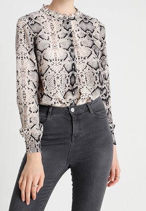 Femme portant un chemisier à manches longues à imprimé serpent, rentré dans un jean skinny gris foncé taille haute, avec des bagues aux doigts.