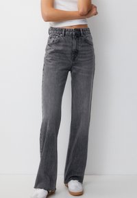Persona con jeans negros desgastados de cintura alta y dobladillos deshechos, de pie contra una pared blanca lisa.