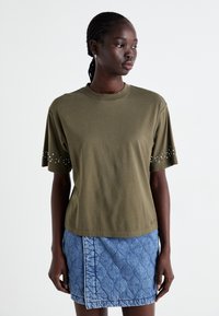 Olijfgroene katoenen t-shirt met korte mouwen, voorzien van zilveren studs langs de mouwzoom, gecombineerd met een blauwe gewatteerde denimrok.