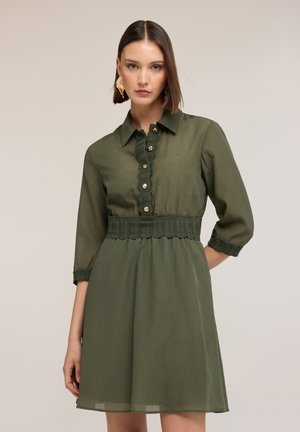 Frau mit glatten braunen Haaren, die ein knielanges olivgrünes Kleid mit Knöpfen, geschwungenem Saum und dreiviertel Ärmeln trägt, steht still.