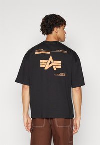 Sort bomulds T-shirt med orange grafisk design, der har "Alpha Industries" og symbol på ryggen. Korte ærmer og afslappet pasform.