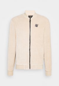 Giacca bomber in corduroy beige con chiusura a zip, colletto e polsini a coste. Presenta un piccolo logo ricamato sul lato sinistro del petto.