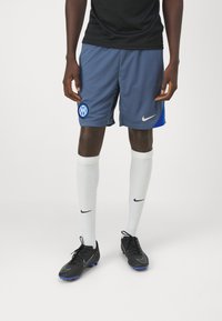 Shorts de sport gris avec des accents bleus, logo Nike et emblème de l'équipe ; associés avec des chaussettes blanches montantes et des crampons noirs avec des semelles bleues.