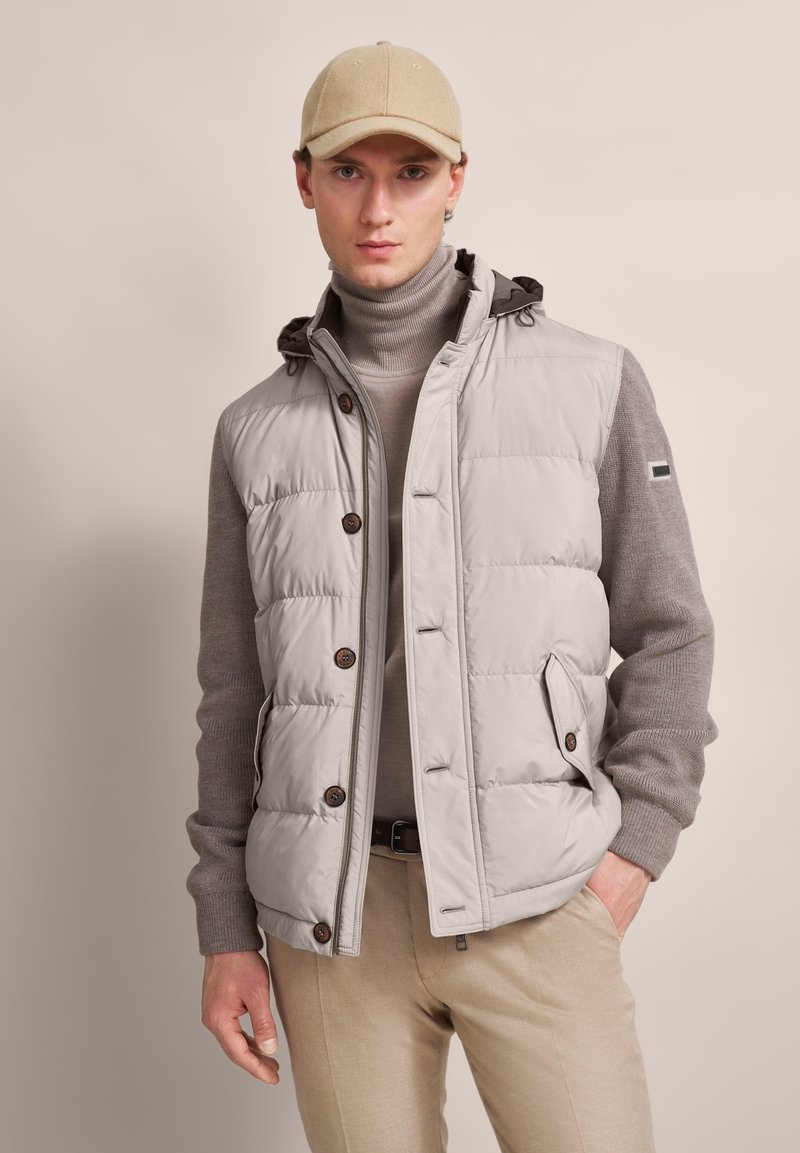 Padded gray vest with brown buttons and a hood over a beige sweater, paired with light-colored pants and a beige cap. Smooth texture.