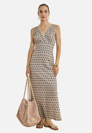 Mujer con vestido largo beige sin mangas con estampado, que lleva un gran bolso de punto y sandalias y pendientes dorados.