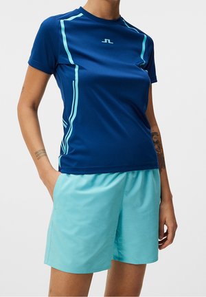 Person trägt ein marineblaues kurzärmeliges Sportshirt mit hellblauen Streifen und hellblauen Shorts, steht mit einer Hand in der Tasche.