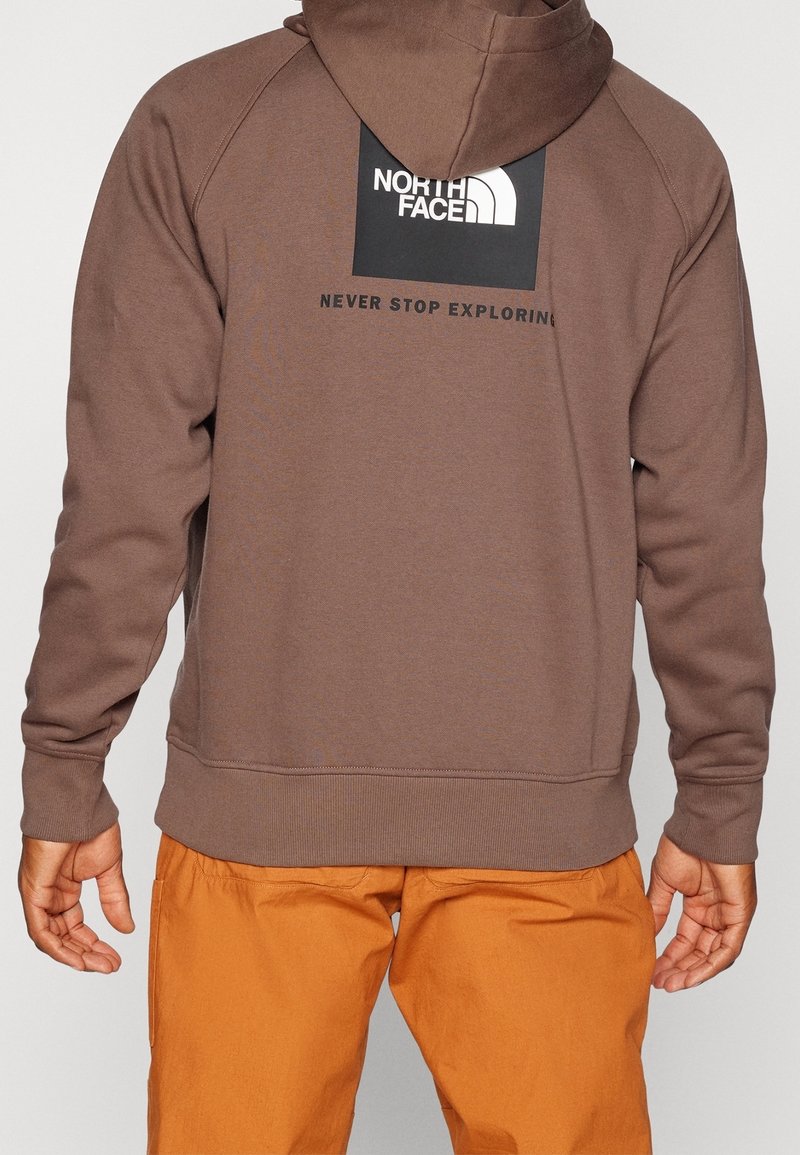 Personne portant un sweat à capuche marron avec un logo noir The North Face et un texte "Never Stop Exploring" dans le dos, assorti à un pantalon orange.