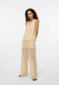 Kanten twee-delige outfit in lichtbeige, met een mouwloze top en brede, semi-transparante broek met ingewikkelde cirkelpatronen.