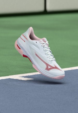 Witte en roze sportschoen zwevend boven de rand van een blauwgroen tennisbaanoppervlak.