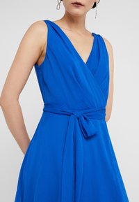 Robe portefeuille bleue avec un profond décolleté en V, sans manches, et une ceinture à nouer à la taille. Le tissu texturé apporte de la profondeur et du mouvement.