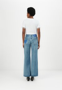 Jeans de denim azul de pierna ancha con cintura alta, con una etiqueta azul en la parte trasera y un diseño de cara sonriente en el bolsillo derecho. Camiseta blanca puesta.
