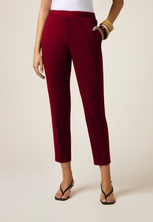 Pantalons taillés bordeaux avec une coupe slim, dotés de poches latérales et d'une texture lisse. Portés avec des sandales flip-flop noires.