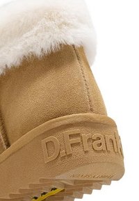 Botín de ante color beige con forro de piel sintética blanca en la parte superior. Cuenta con una suela de goma con "D.Frank" grabado en el lateral y detalles en amarillo.
